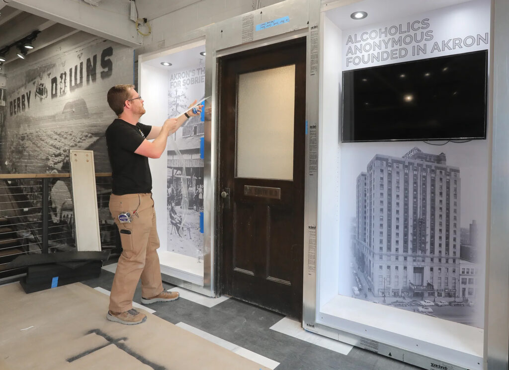 Mike Hill, a production supervisor from CEl, removes a protective coating Thursday from an Alcoholics Anonymous exhibit at the Akron History Center. In the middle is A.A. co founder Dr. Bob Smith's office door. The building in the vintage photo at right is the Mayflower Hotel. PHIL MASTURZO, AKRON BEACON JOURNAL