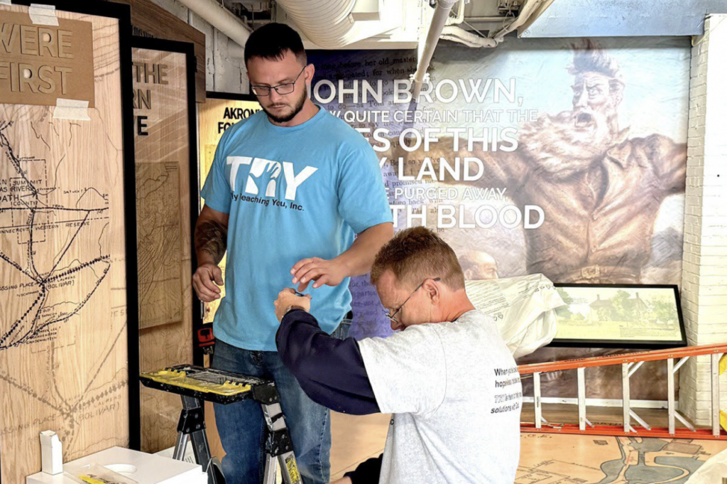 Jamie Rhodes (left) and Aaron Engstrom add lettering to an exhibit in the Akron History Center building in downtown Akron, Oct. 24, 2024. The two volunteers are with Truly Reaching You, Inc., an Akron-based ministry helping men with re-entry after incarceration. (Kevin Dilley / Signal Akron)